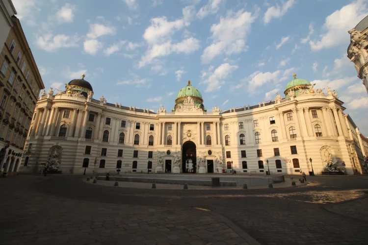 eine Panoramaaufnahme der Wiener Hofburg, das barocke Gebäude ist von der Sonne erleuchtet, davor liegt der Michaelerplatz im Schatten.