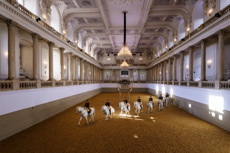 mehrere Pferde reiten hintereinander mit ihren Bereiter:innen in barocker Reithalle