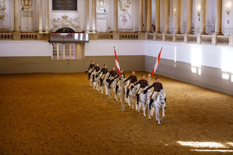 eine Reihe von Pferden und Reitern reiten durch Pilaren in einer barocken Reithalle, sie ziehen ihren Zweispitz und grüßen