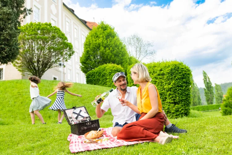 zwei Erwachsene picknicken auf einer grünen Wiese, im Hintergrund spielen zwei Mädchen